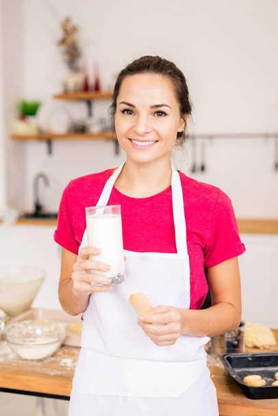 Happy young brunette female with toothy smile having glass of milk with baked homemade cookie in front of camera - Foto, Imagem