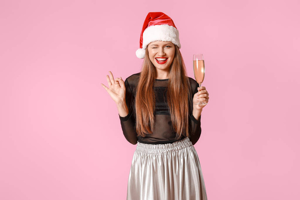 Happy young woman in Santa Claus hat and with glass of champagne showing OK gesture on color background - Foto, afbeelding