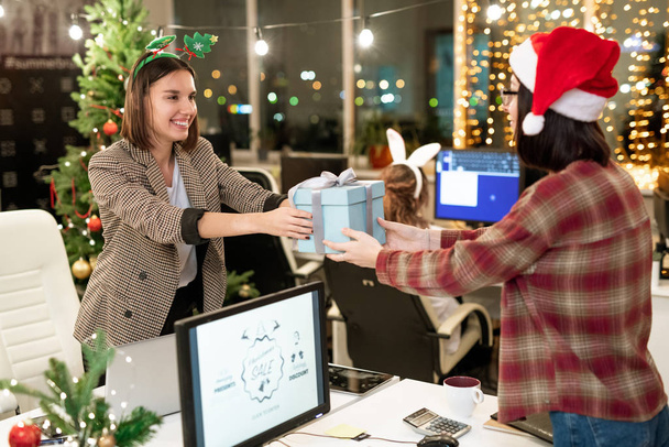 Two happy young businesswomen giving presents to each other over computer monitor on Christmas day in office - 写真・画像