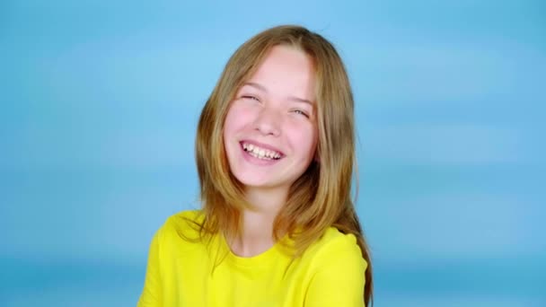 Η χαρούμενη έφηβη με το κίτρινο μπλουζάκι γελάει και κοιτάζει την κάμερα. Μπλε φόντο με χώρο αντιγραφής. Εφηβικά συναισθήματα. 4k πλάνα - Πλάνα, βίντεο