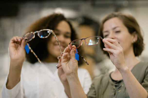 Dłonie młodej konsultantki i jej klientki porównujące dwa modele okularów z nowej kolekcji przy jednoczesnym omówieniu ich cech - Zdjęcie, obraz
