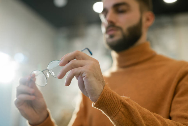 Руки молодого чоловіка в жовтому пуловері, що тримає пару нових окулярів при виборі окулярів в магазині оптики
 - Фото, зображення