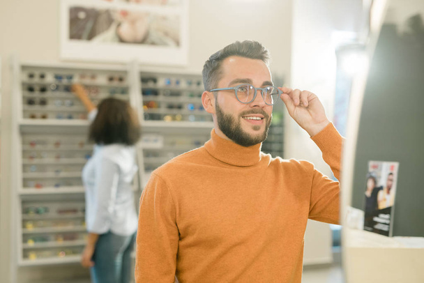 Mladý usměvavý muž ve svetru zkouší nové brýle před zrcadlem na pozadí ženského kupce na displeji s brýlemi - Fotografie, Obrázek