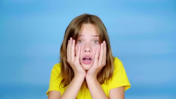 Sarı tişörtlü genç kız ellerini kafasına koyar, ağzını açar ve çok şaşırır. Kopyalama alanı olan mavi arkaplan. Gençlik duyguları. 4k görüntü - Video, Çekim