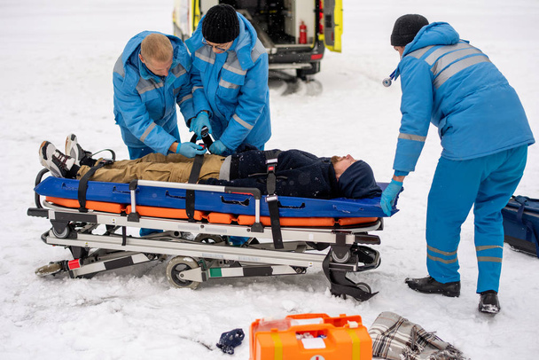 Két fiatal mentős kék munkaruhában megjavítja az eszméletlen férfit övekkel a hordágyon, mielőtt a mentőautóba szállítják. - Fotó, kép