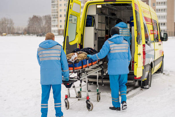 Dois jovens paramédicos em azul workwear empurrando maca com homem inconsciente fixo em carro de ambulância
 - Foto, Imagem