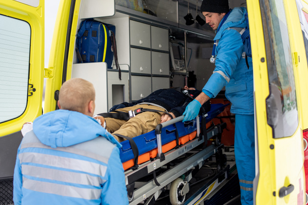 Paramedics in uniform pushing stretcher with fixed unconscious man into ambulance car to get him to hospital - Fotoğraf, Görsel