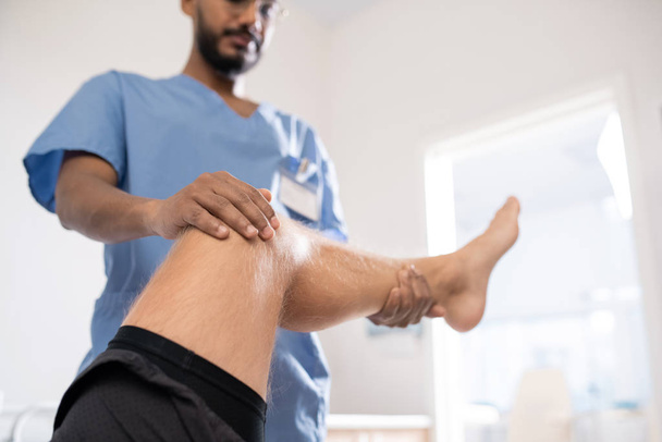 Joven médico contemporáneo de raza mixta sosteniendo pierna enferma de su paciente mientras lo examina en el hospital
 - Foto, Imagen