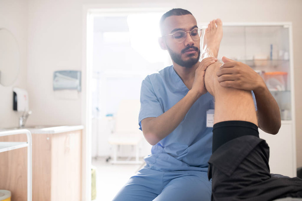 Młody profesjonalny masażysta trzymający chorą nogę pacjenta na ramieniu podczas masażu kolana w klinikach - Zdjęcie, obraz