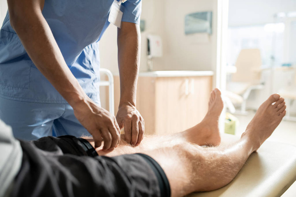 Zieke patiënt liggend op de bank terwijl clinicus in blauw uniform voorover buigt en zijn been masseert - Foto, afbeelding