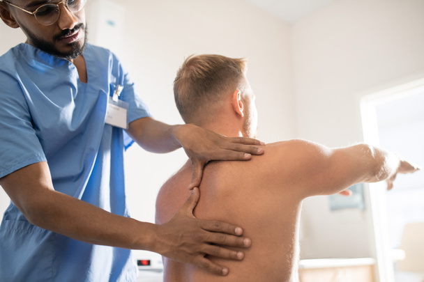 Young medical specialist in uniform massaging shoulder and back of shirtless patient in clinics - 写真・画像