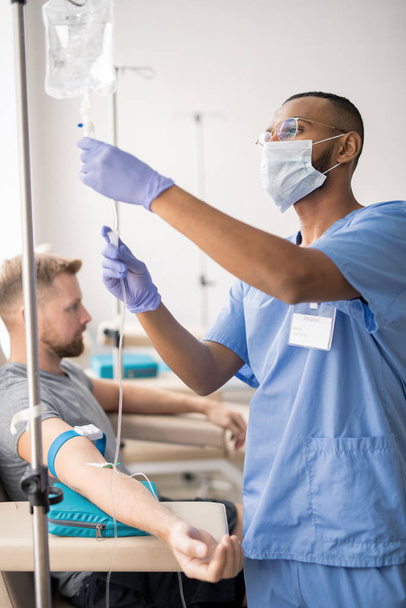 Молодий лікар змішаної раси в блакитній формі, захисна маска і гумові рукавички готують крапельницю для одного з пацієнтів, що сидять поруч
 - Фото, зображення
