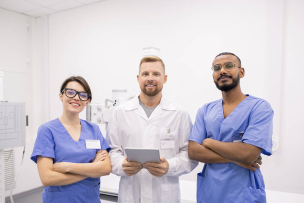 Grupo de tres jóvenes y exitosos médicos interculturales o pasantes uniformados de pie frente a la cámara en el hospital
 - Foto, imagen