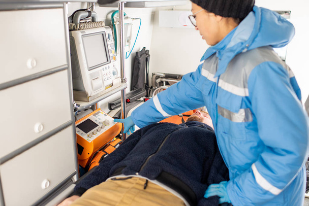 Junge Sanitäter in Uniform drücken Taste an medizinischem Erste-Hilfe-Gerät, während sie bewusstlosen Mann auf Trage stehen - Foto, Bild