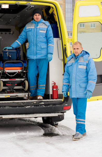 One of male paramedics in uniform going to take stretcher while his colleague standing by ambulance car - 写真・画像