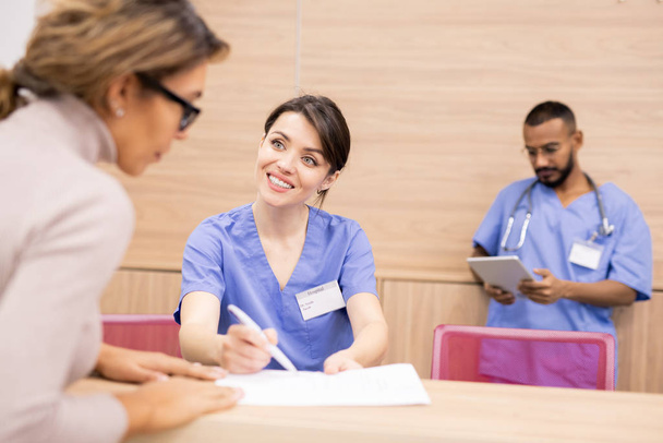 hübsche Ärztin in Uniform lächelt Patientin an, während sie auf ärztliches Dokument zeigt und junge Frau berät - Foto, Bild