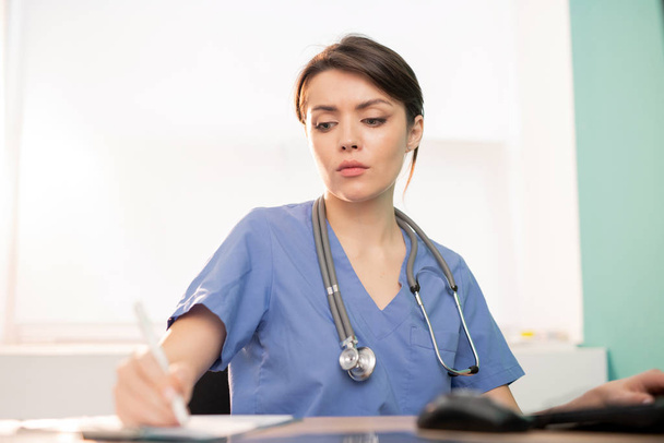 Giovane medico serio o stagista in uniforme blu prendere appunti di lavoro mentre seduto sul posto di lavoro di fronte al computer
 - Foto, immagini