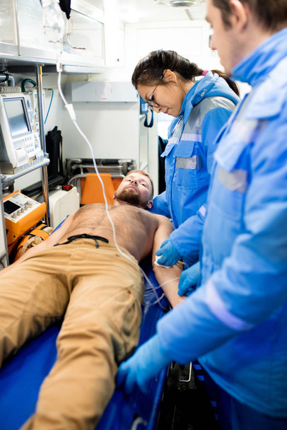Young female paramedic in uniform giving first aid to sick shirtless man lying on stretcher inside ambulance car - 写真・画像