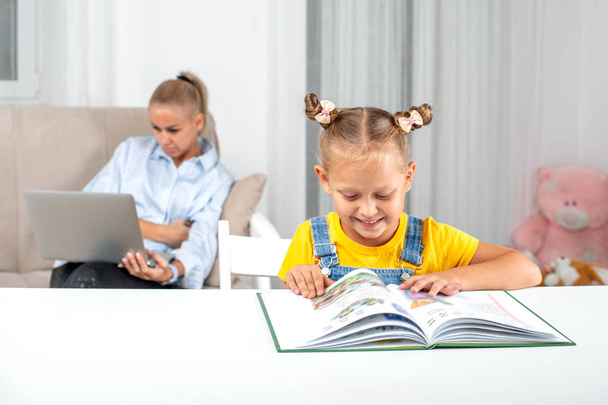Shot of a smiling girl sitting at the table at homeand reading b - Photo, Image