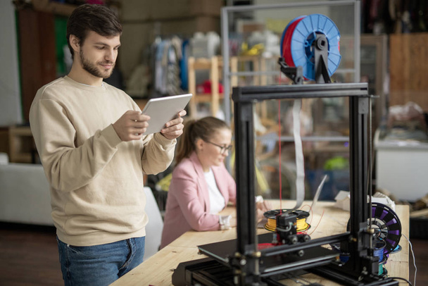 Young man with touchpad looking at online figure on display while standing in front of 3d printer on background of colleague - Foto, imagen