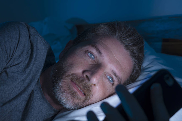 mladý atraktivní a uvolněný muž s modrýma očima ležící na posteli pozdě v noci v tmavé a matné světlo sítí na mobilním telefonu nebo online randění v závislosti na internetu  - Fotografie, Obrázek