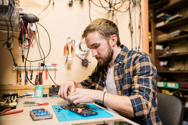 Профессиональный ремонтник, наклоняющийся над сломанным смартфоном и исправляющий мелкие детали во время работы за столом в мастерской
 - Фото, изображение