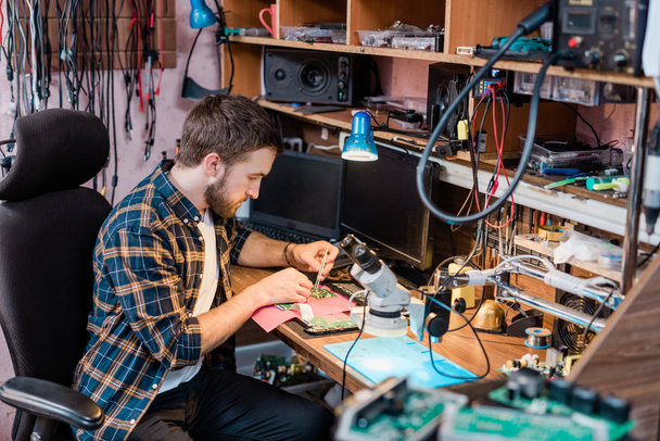 Mladý profesionální mechanik s malými ocelovými pinzetami opravující demontovaný touchpad nebo jiný přístroj ve své dílně - Fotografie, Obrázek
