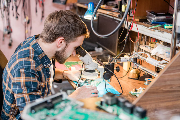 Επαγγελματίας επισκευαστής gadget ψάχνει στο μικροσκόπιο ανά χώρο εργασίας, ενώ προσπαθεί να βρει το πρόβλημα του smartphone ή tablet - Φωτογραφία, εικόνα