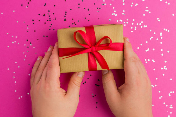 Man hands holding Brown gift boxes with red ribbons. Pink background with multicolored confetti. Flat lay style. Gift for Christmas, valentine day, mother day or a birthday. - Fotografie, Obrázek