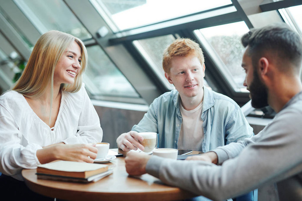 Grupa szczęśliwych przyjaciół z koledżu siedzących przy stole w kawiarni po zajęciach, pijących kawę i omawiających plany na tydzień - Zdjęcie, obraz