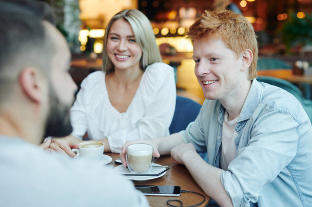 Happy casual guy with cup of cappuccino and his girlfriend looking at their friend during chat by table in cafe - Photo, Image