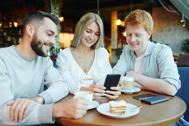 Pretty blonde girl with smartphone showing two happy guys her new photos while relaxing by cup of cappuccino by table in cafe - Foto, Imagem