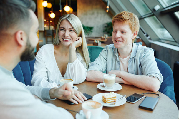 Heureux jeune couple occasionnel bavarder avec leur ami par tasse de cappuccino et dessert tout en étant assis à table dans un café
 - Photo, image