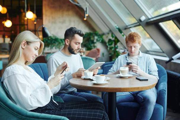 hübsches blondes Mädchen und zwei Jungs sitzen in blauen weichen Samtsesseln an einem Tisch im Café und bedienen sich in ihrer Freizeit mobiler Geräte - Foto, Bild
