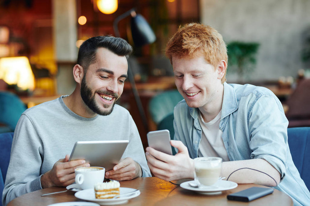 Два счастливых парня с мобильными гаджетами смотрят любопытные вещи в смартфоне одного из них, проводя время в кафе
 - Фото, изображение