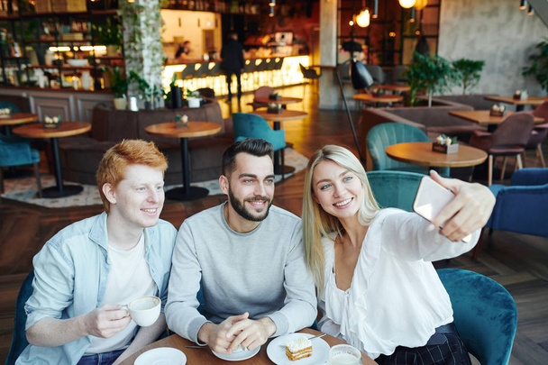 Tres jóvenes amigos felices sentados a la mesa en el acogedor café, haciendo selfie y disfrutando del café con postre
 - Foto, Imagen