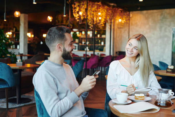 Happy blonde girl with copybook looking at young man with smartphone while spending time by cup of tea in cafe - Photo, Image