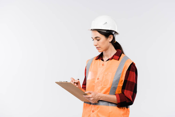Young female builder in hardhat, flannel and working vest making notes in document in front of camera in isolation - Zdjęcie, obraz