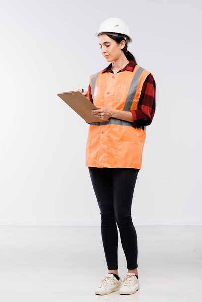 Pretty female builder in hardhat, flannel, leggins and working vest making notes in document in front of camera in isolation - Photo, Image