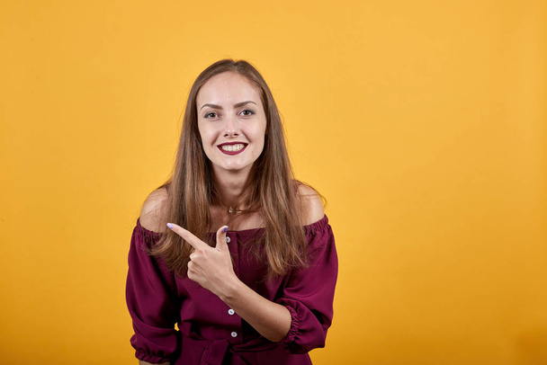 junge Dame in weinroter Bluse über vereinzelter orangefarbener Wand zeigt mit dem Finger nach links - Foto, Bild