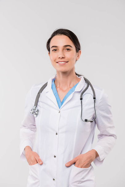 jong glimlachen brunette clinicus in whitecoat houden haar handen in zakken terwijl staan in de voorkant van de camera in isolatie - Foto, afbeelding