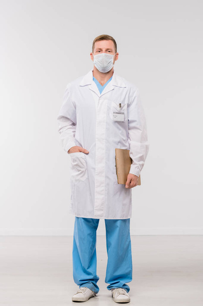 Nuori lääkäri tai kirurgi valkotakissa, yhtenäinen ja suojaava naamio tilalla asiakirjan seistessään eristyksissä
 - Valokuva, kuva