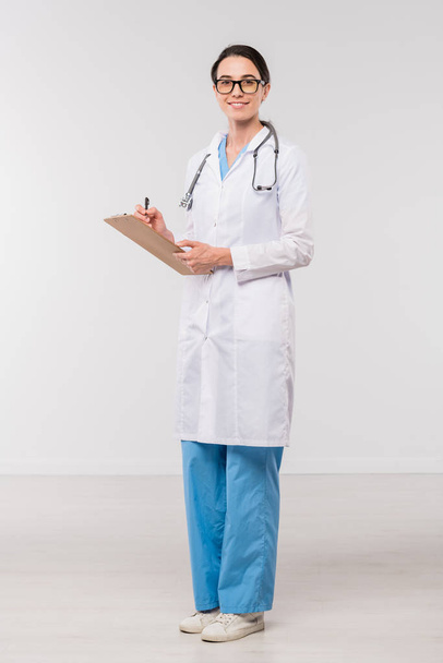 Young confident clinician in whitecoat filling in medical document while standing in front of camera in isolation - Photo, Image
