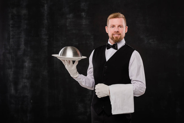Camarero elegante feliz de restaurante con clase sosteniendo toalla blanca y cloche con comida mientras está de pie sobre fondo negro
 - Foto, Imagen