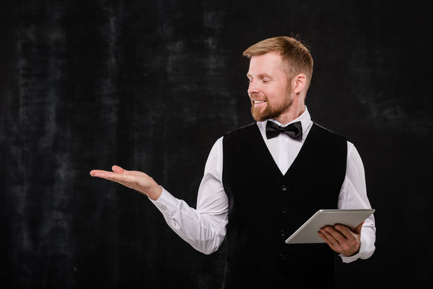 Young elegant waiter with touchpad looking at imaginary stuff on his hand while standing in front of camera against black background - Photo, Image