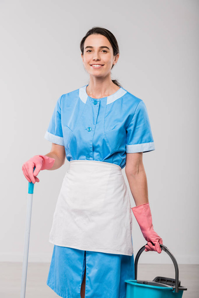 Happy young hotel room cleaning staff in uniform and rubber gloves holding bucket and mop while standing in front of camera in isolation - Photo, Image