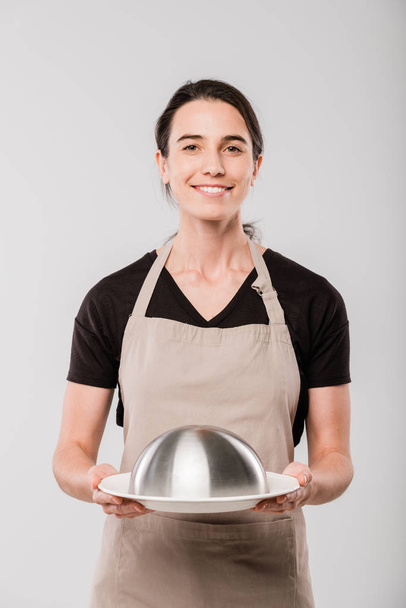 Ευτυχισμένη νεαρή μελαχρινή σερβιτόρα με ποδιά κρατώντας πανιά με έτοιμο φαγητό για τον πελάτη, ενώ στέκεται μπροστά από την κάμερα στην απομόνωση - Φωτογραφία, εικόνα