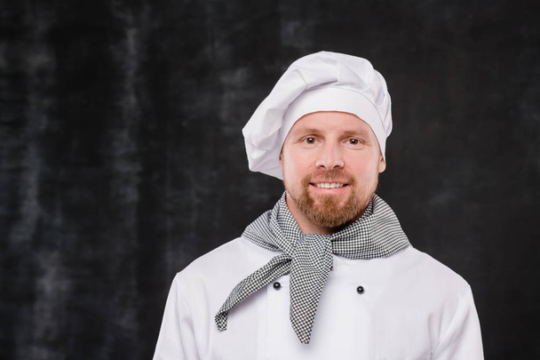 Chef barbudo feliz en uniforme blanco mirando a la cámara con sonrisa mientras está de pie sobre fondo negro
 - Foto, Imagen