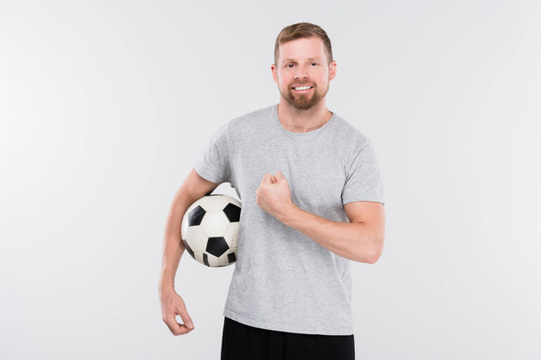Νεαρός επιτυχημένος ποδοσφαιριστής στα αθλητικά κρατώντας μπάλα και δείχνοντας τη δύναμή του μπροστά στην κάμερα στην απομόνωση - Φωτογραφία, εικόνα