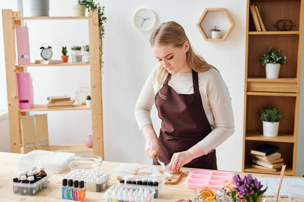 Νεαρή δημιουργική ξανθιά γυναίκα σε καφέ ποδιά κοπής σαπουνιού μάζα σε ξύλινο πίνακα, ενώ εργάζονται σε στούντιο - Φωτογραφία, εικόνα